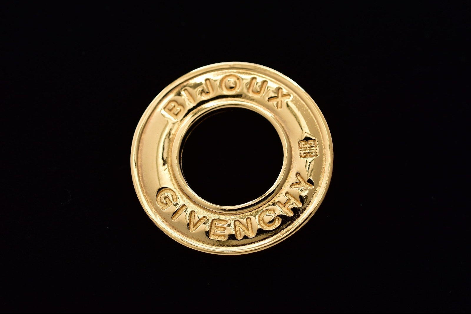 A circular gold-tone metal ring charm featuring the embossed brand names 'BIJOUX' and 'GIVENCHY' on a solid black background.