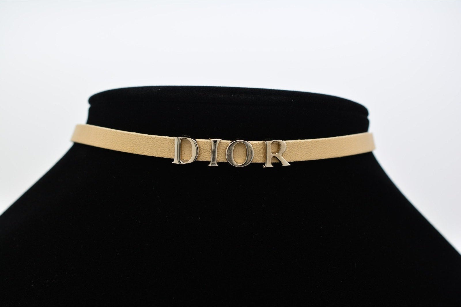 A Dior belt in beige leather with a gold-tone metal logo bar spelling 'DIOR', displayed on a black velvet jewelry bust against a white background.
