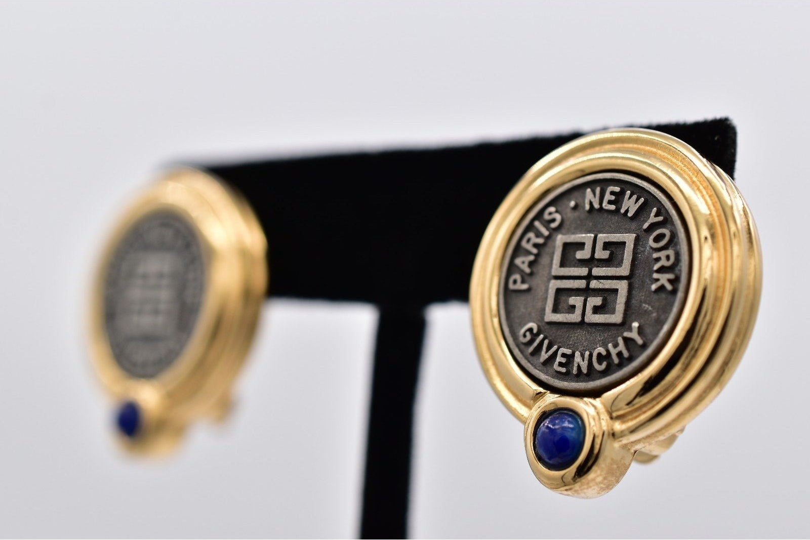 A pair of gold-toned Givenchy cufflinks displayed on a black T-stand against a light gray background. One cufflink is in sharp focus, showing a circular black face with the Givenchy logo, 'PARIS - NEW YORK' text, and a small blue gemstone accent.