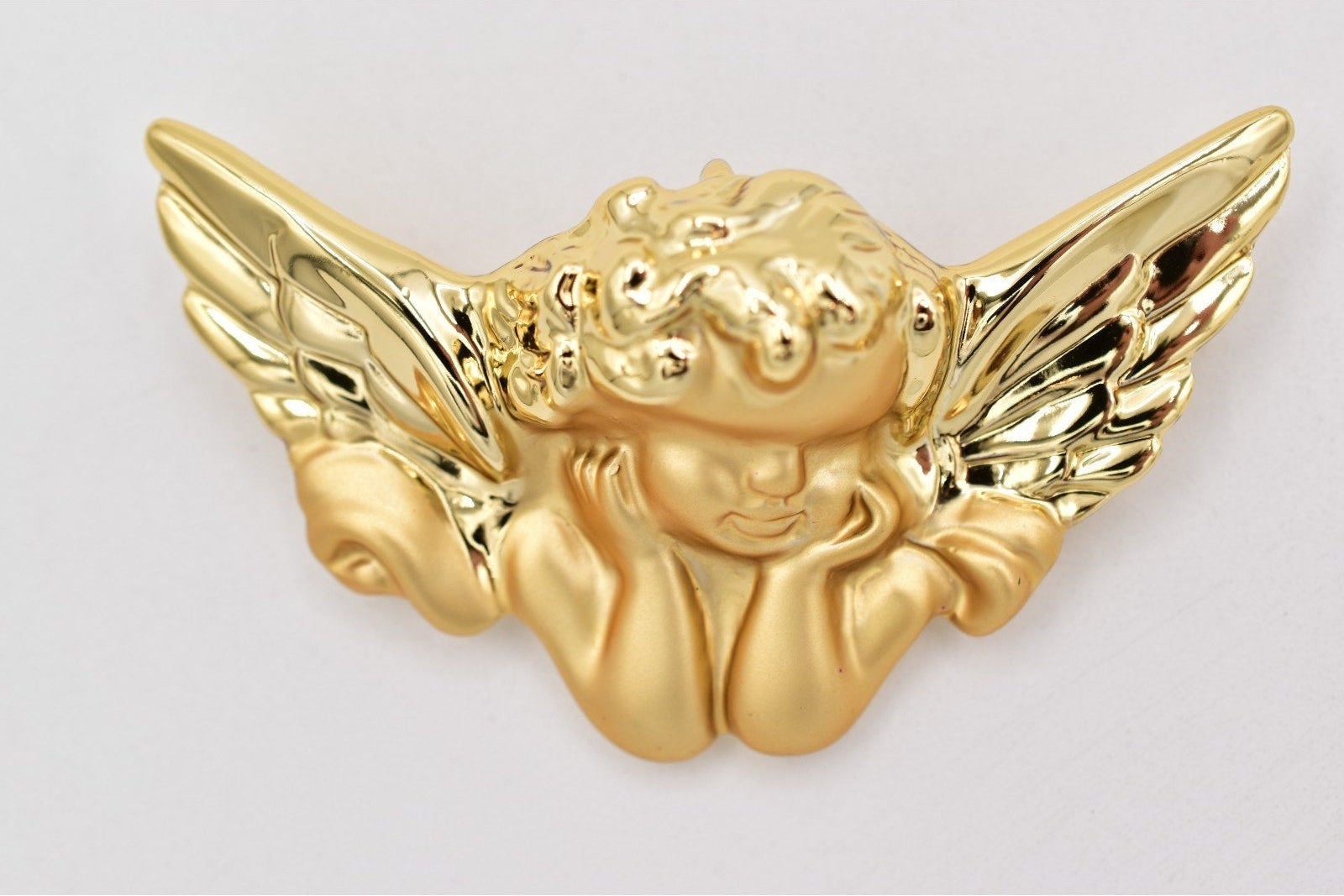 A polished gold cherub wing emblem or badge, featuring a kneeling cherub with detailed wings, shown on a light gray background.