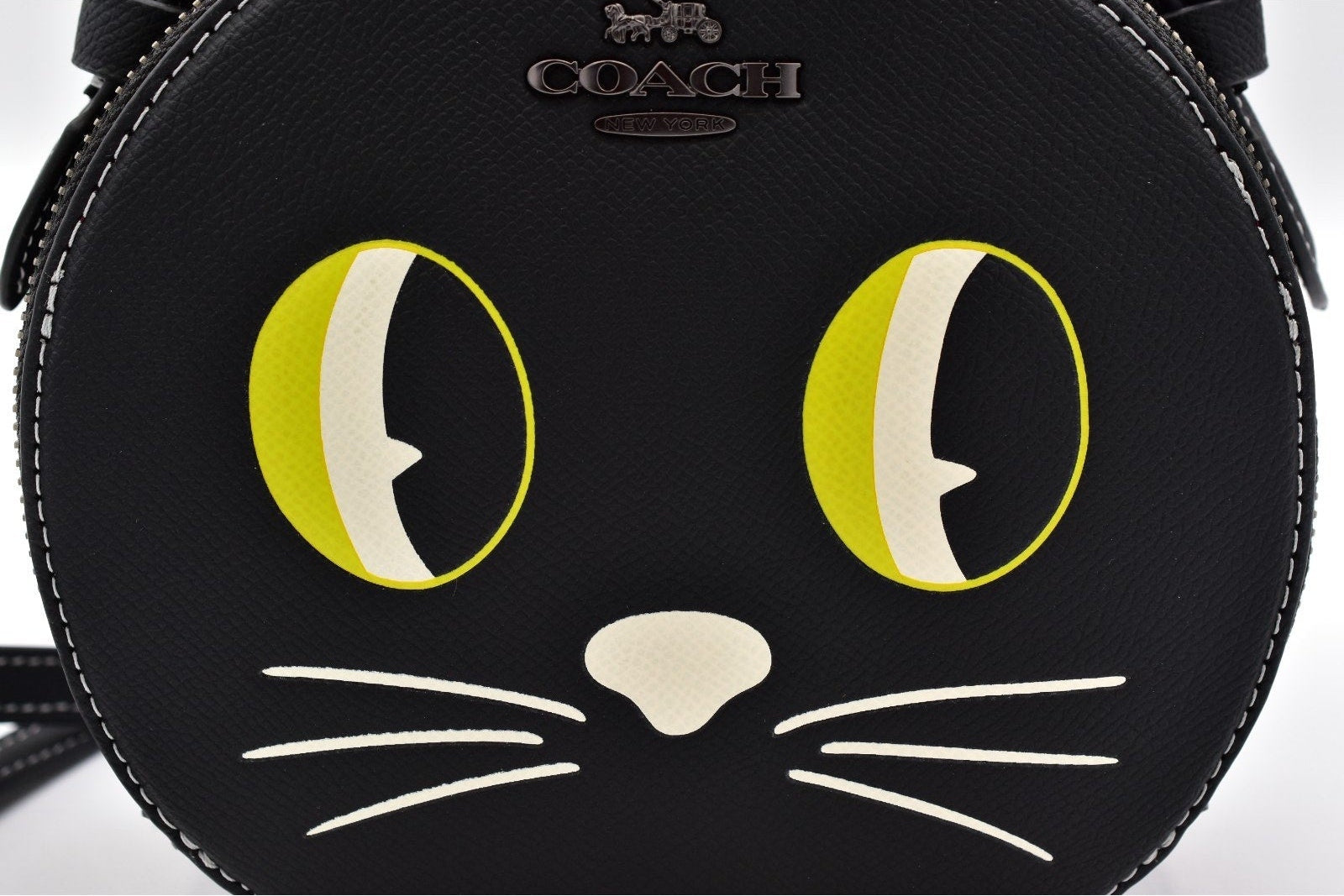 A close-up of a black leather Coach cat-face crossbody bag featuring large yellow and white eyes, a cream-colored nose, and white whiskers, with the Coach logo embossed above.