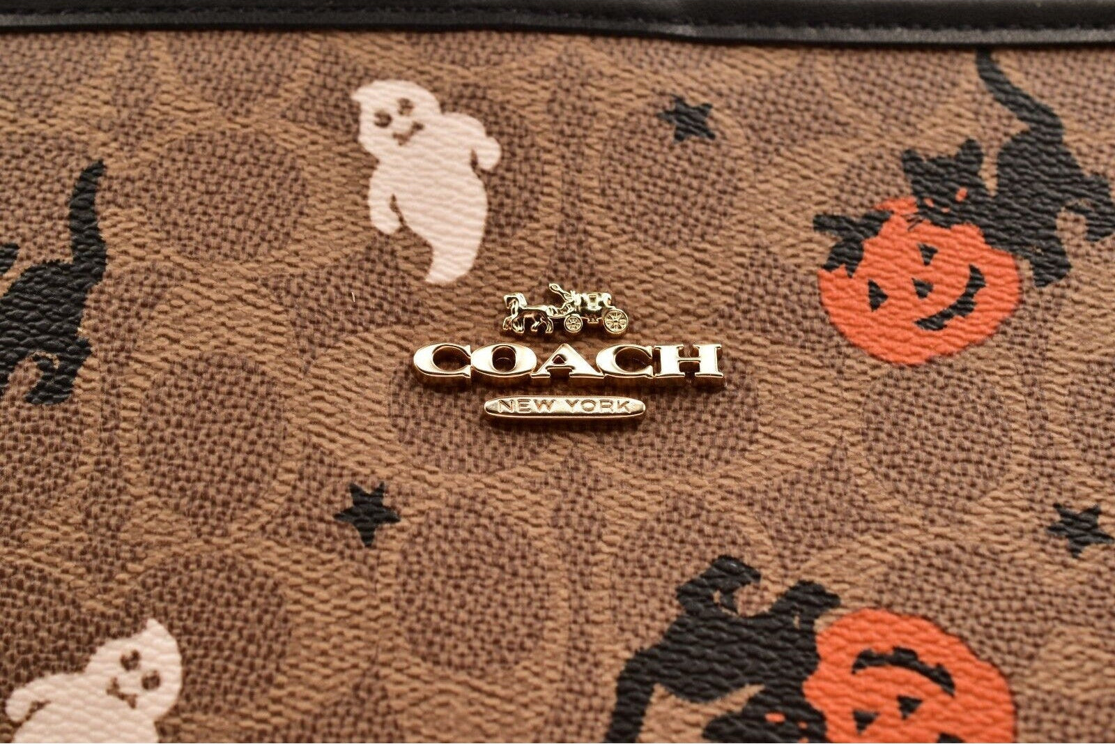 Close-up of a Coach New York wallet featuring the brand's signature brown monogram pattern with Halloween motifs including black cats, white ghosts, and orange jack-o'-lanterns, with a gold-tone COACH logo plaque at the center.