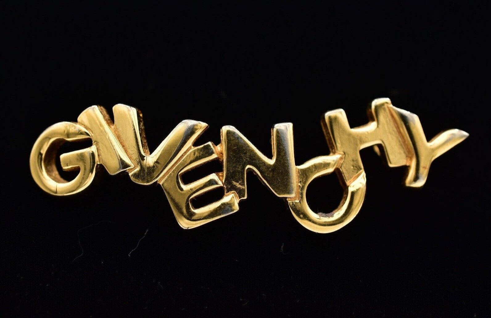 A vintage Givenchy gold logo brooch with block letters on a black background.