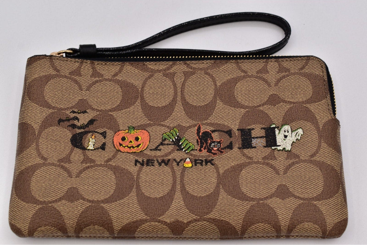 A Coach New York wristlet pouch in the signature brown monogram canvas, featuring embroidered 'COACH' lettering with Halloween-themed embellishments (a pumpkin, spider, cat, and ghost) and a black wrist strap.