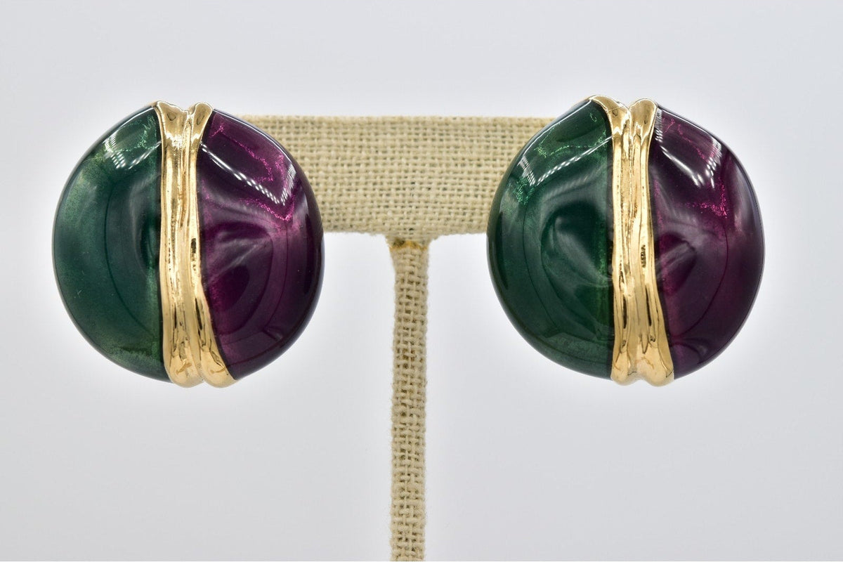 A pair of round stud earrings with polished gold-toned metal and split green and purple stone inlays, displayed on a beige jewelry stand against a light gray background.