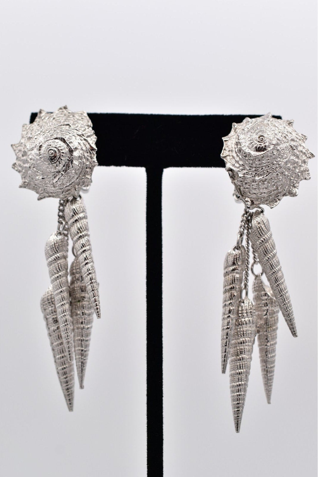 Pair of silver shell and spike earrings on a black stand against a white background