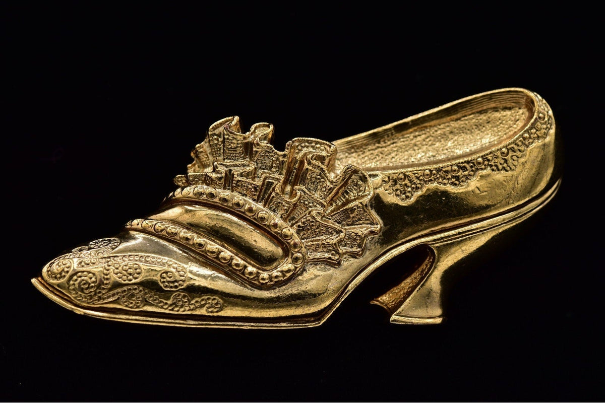 An ornate gold-colored high-heeled shoe with intricate embossed detailing and a decorative strap across the instep, shown against a solid black background.