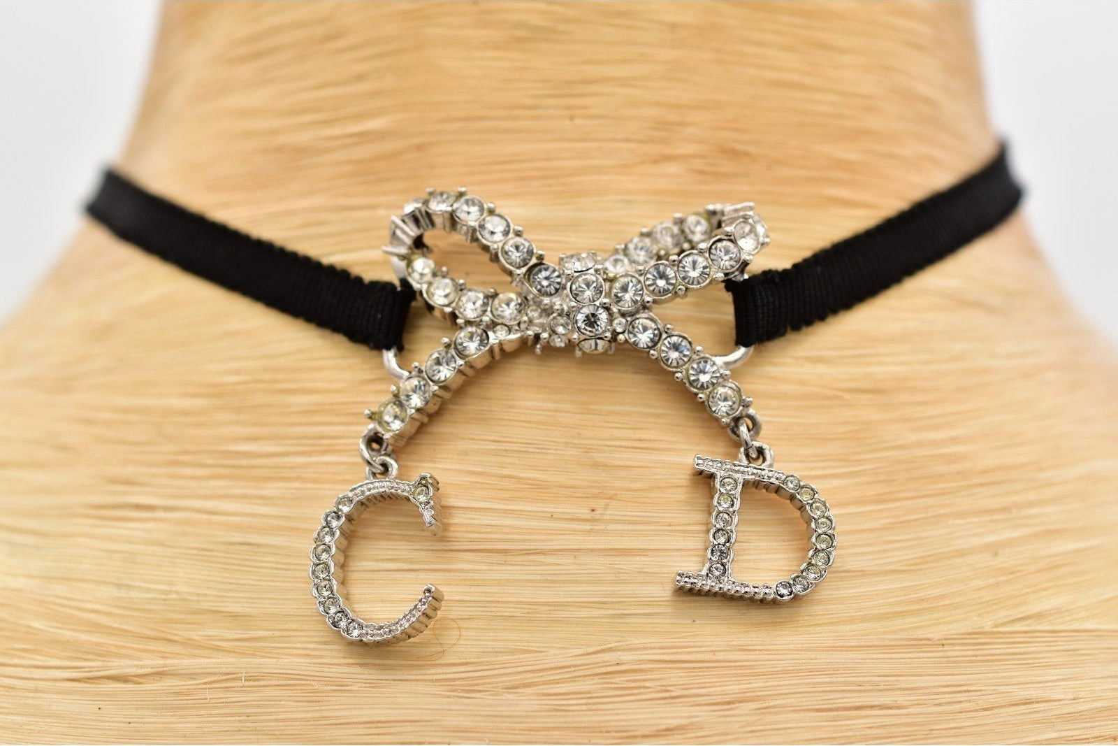 A black ribbon choker necklace displayed on a light wood mannequin neck, featuring a sparkling silver crystal-embellished bow at the center with dangling letter charms 'C' and 'D' set with clear rhinestones.