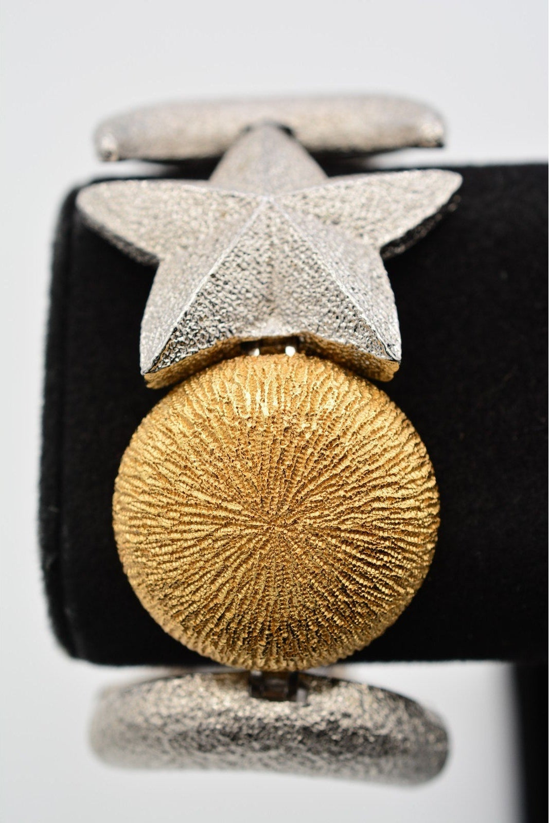 A close-up of a textured gold and silver cufflink on a black display stand against a light gray background.