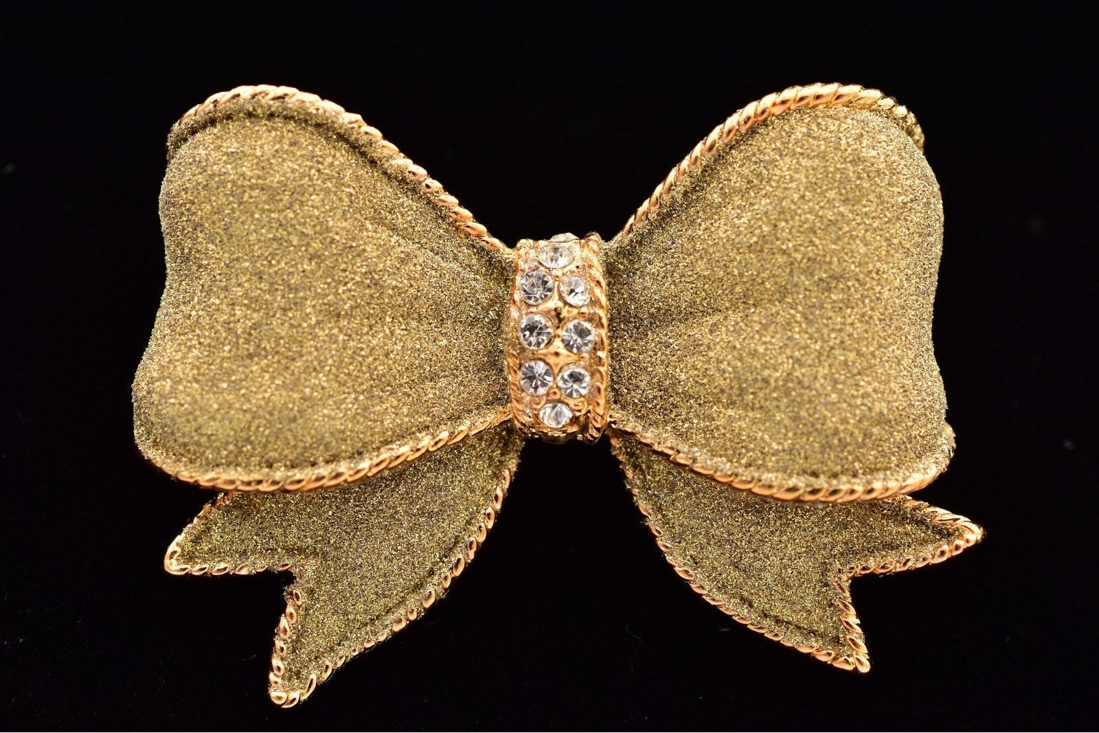 A gold bow-shaped brooch with a textured glitter finish, a central band of clear rhinestones, and a braided gold border, shown on a solid black background.