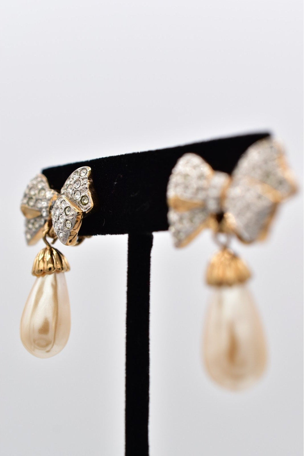 Gold bow-shaped earrings with pearl drops on a black stand against a white background