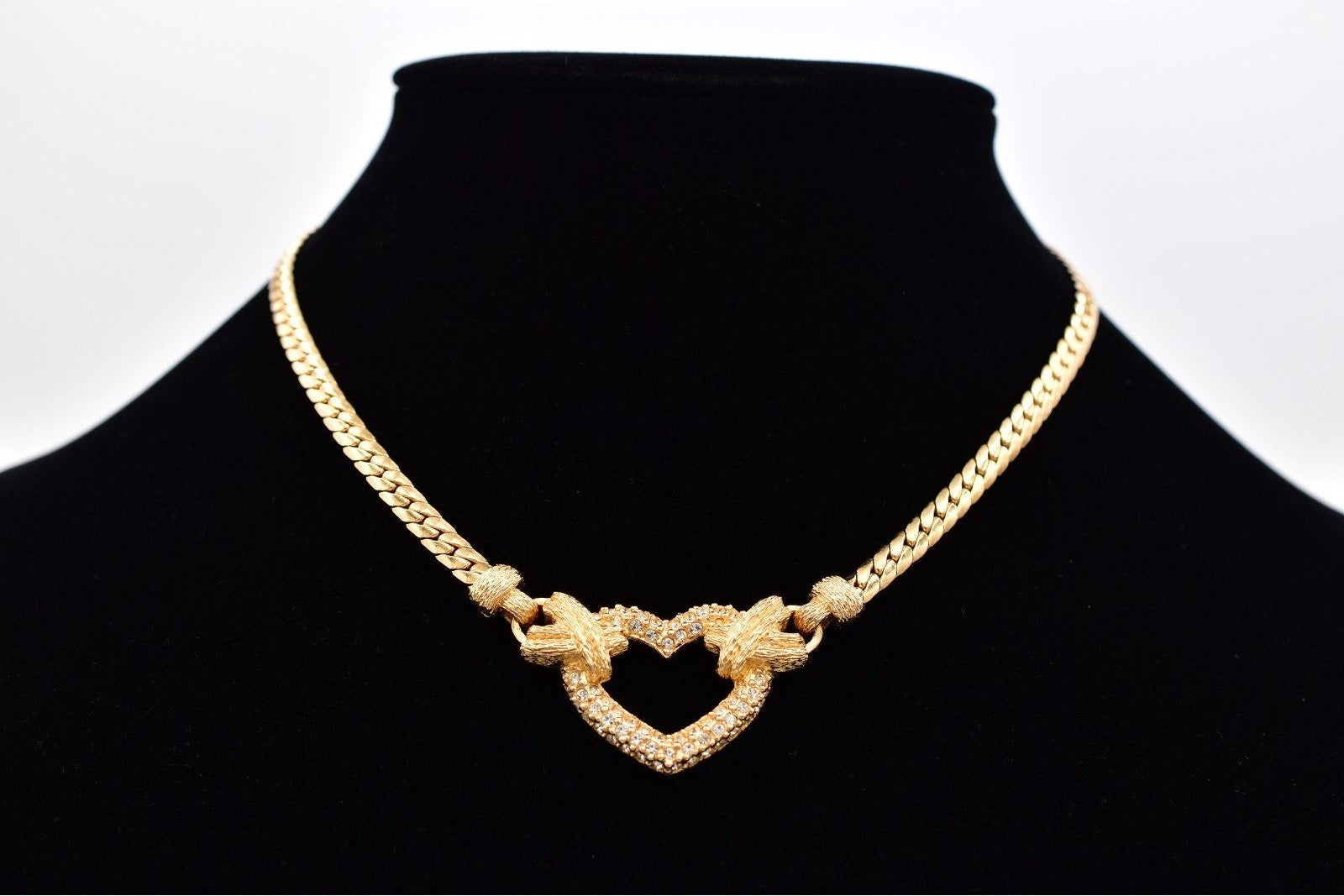A gold-toned chain necklace with a heart-shaped pendant, displayed on a black jewelry bust against a white background.