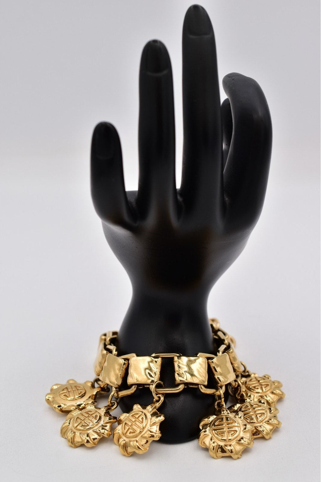 A gold-tone charm bracelet displayed on a black hand-shaped mannequin against a light gray studio background.