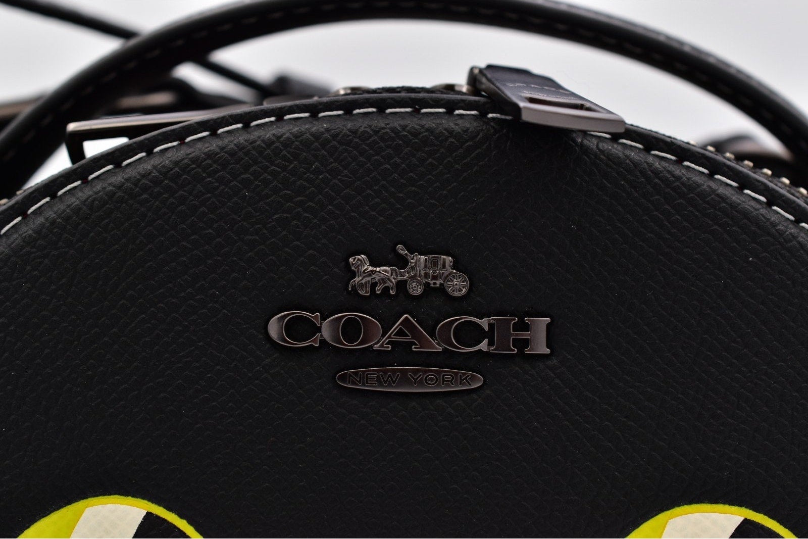 Close-up of a black textured leather Coach handbag with a silver-tone metal logo plaque featuring the horse and carriage emblem and the brand name 'COACH'.