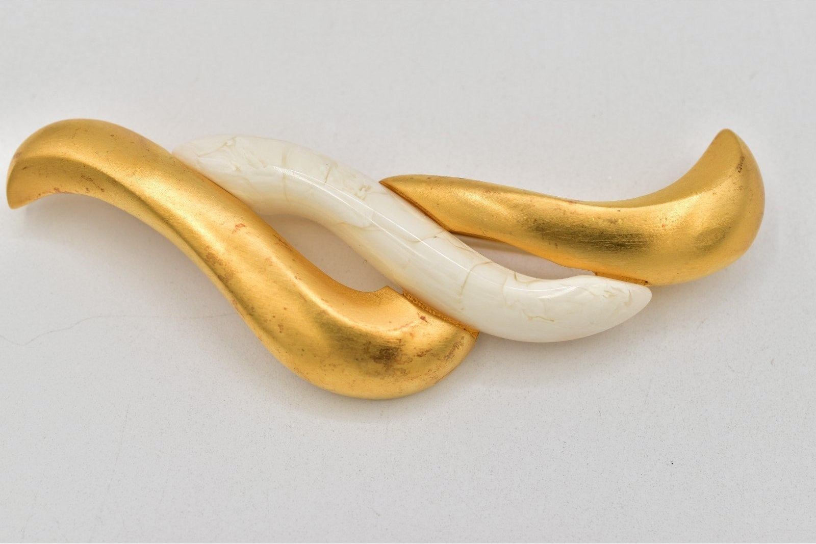 A modern abstract wavy brooch featuring two intertwined fluid shapes, one in polished gold-tone metal and the other in smooth off-white stone or resin.