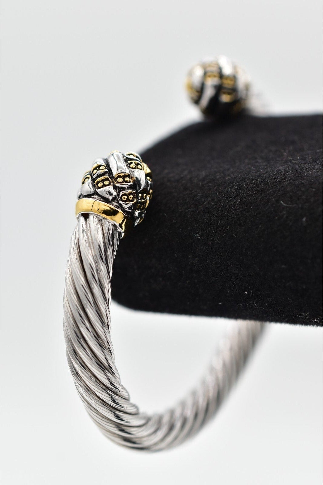 Silver cable-style bracelet with gold accents on a black stand against a white background
