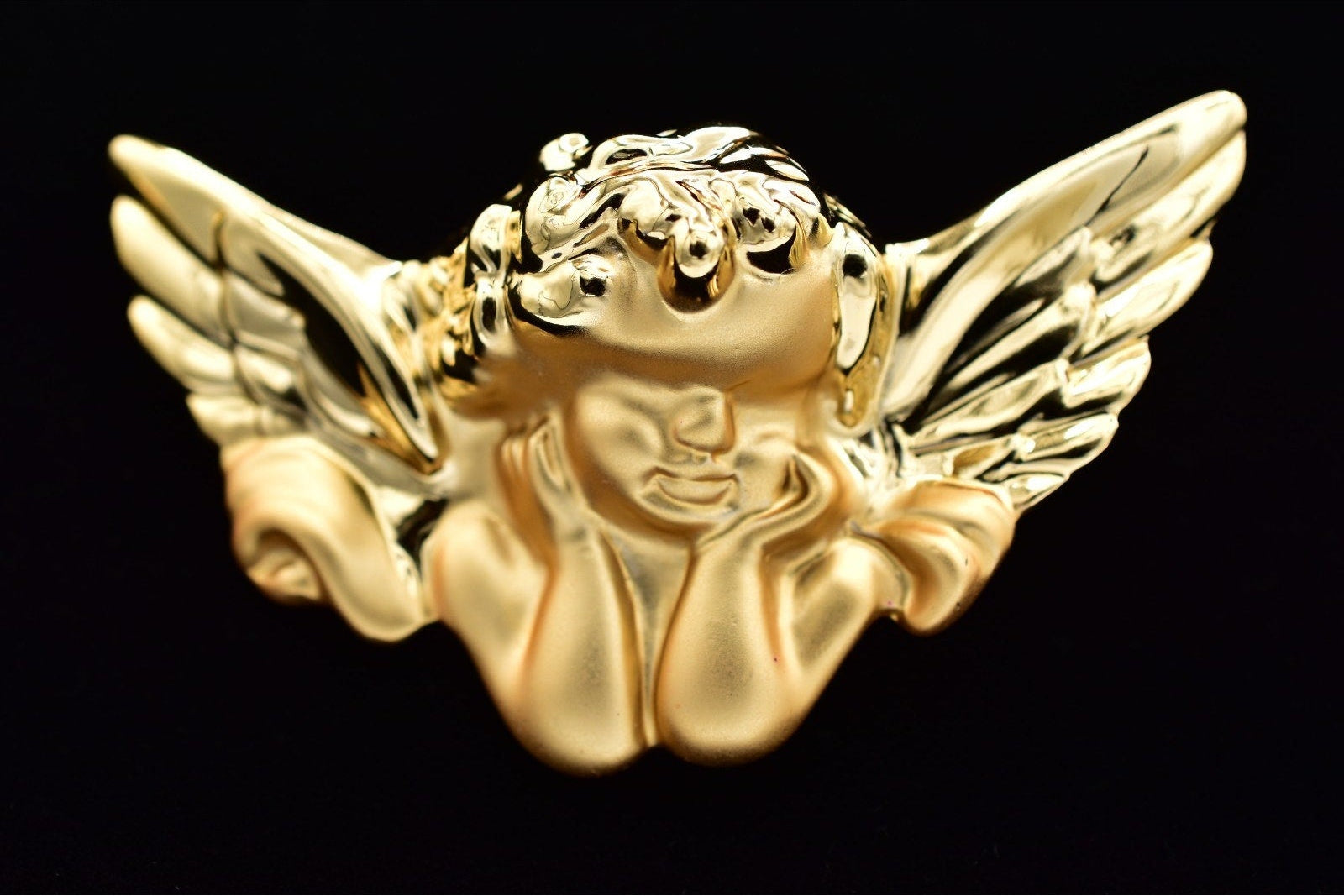 A shiny gold cherub winged bust brooch or pin, shown against a black background.