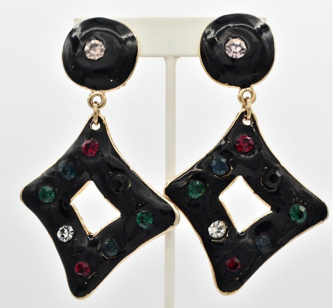View of a pair of vintage black enamel clip on earrings with red, blue, green, black, and clear rhinestone crystals hanging on a white display stand