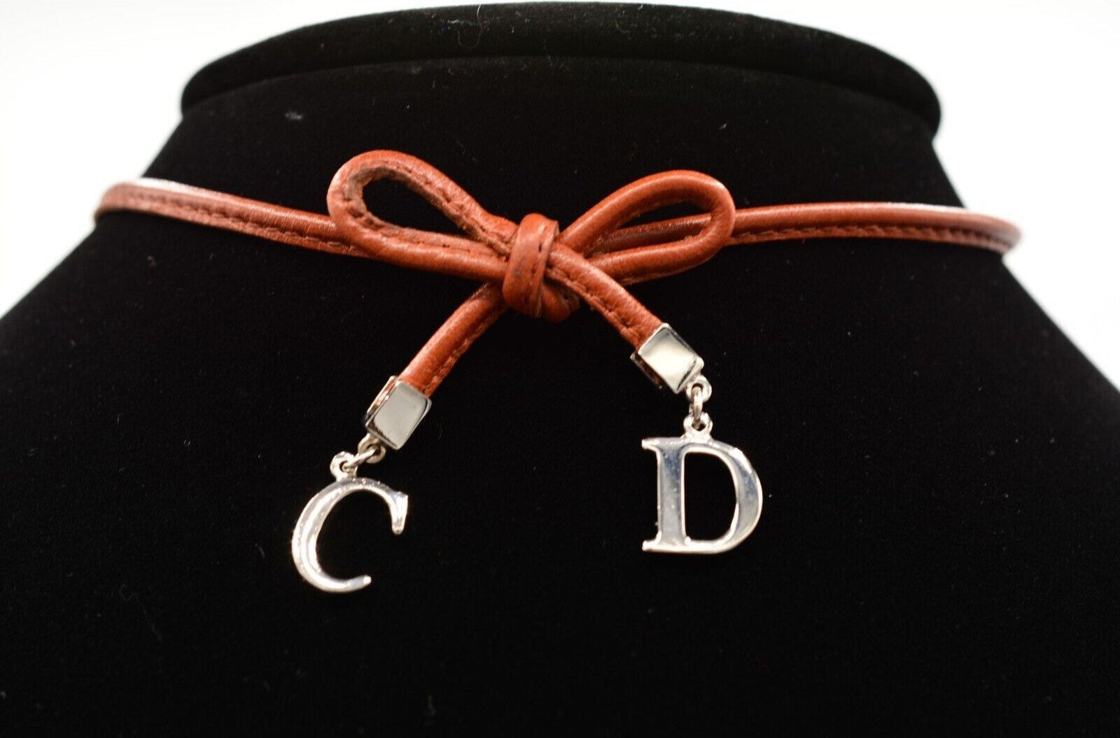 View of the front of a vintage brown leather Christian Dior choker necklace with a bow tied in the middle and dangling silver CD charms on a black velvet background