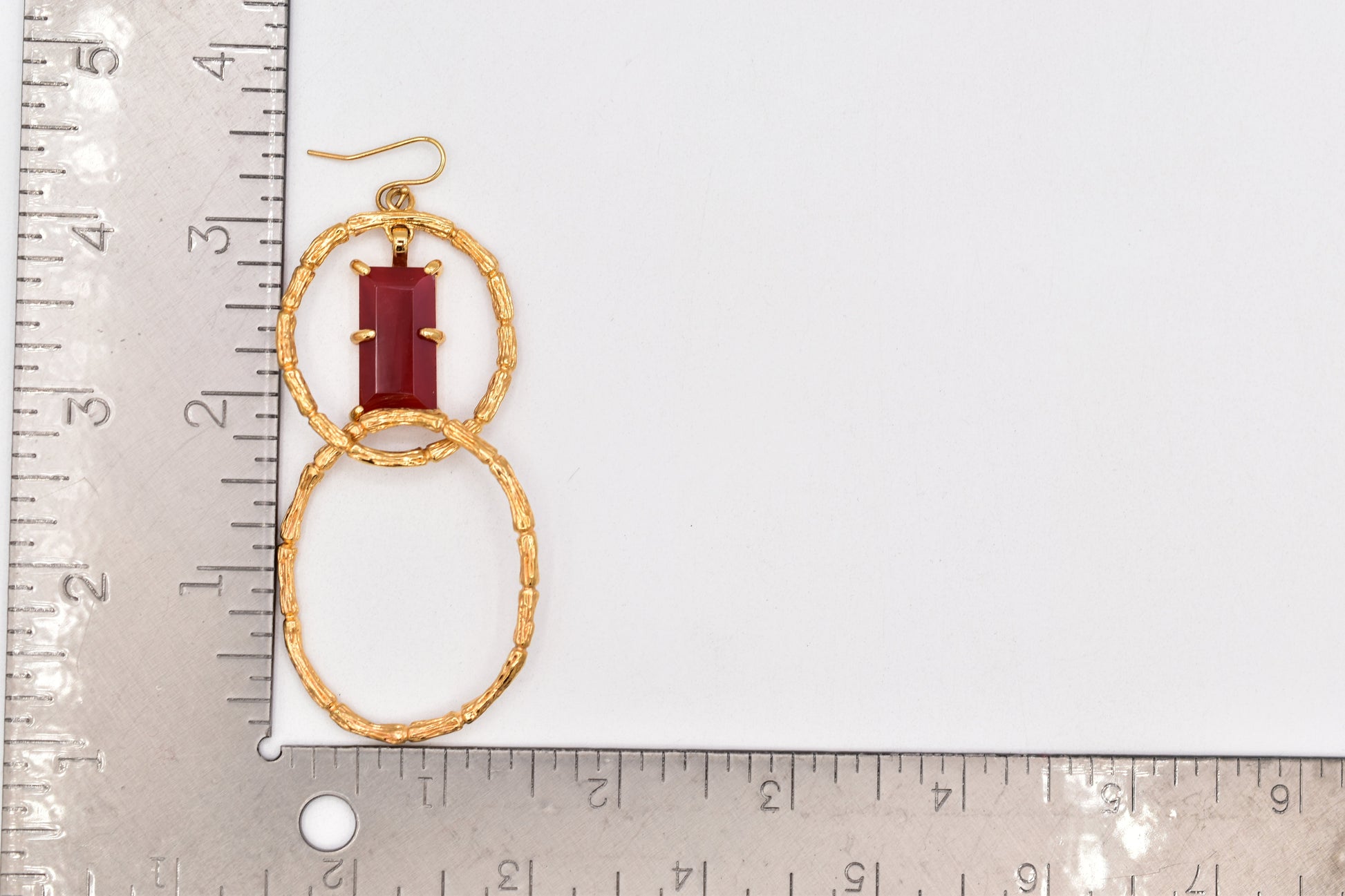 Gold earrings with a red gemstone on a white background with a ruler for scale.