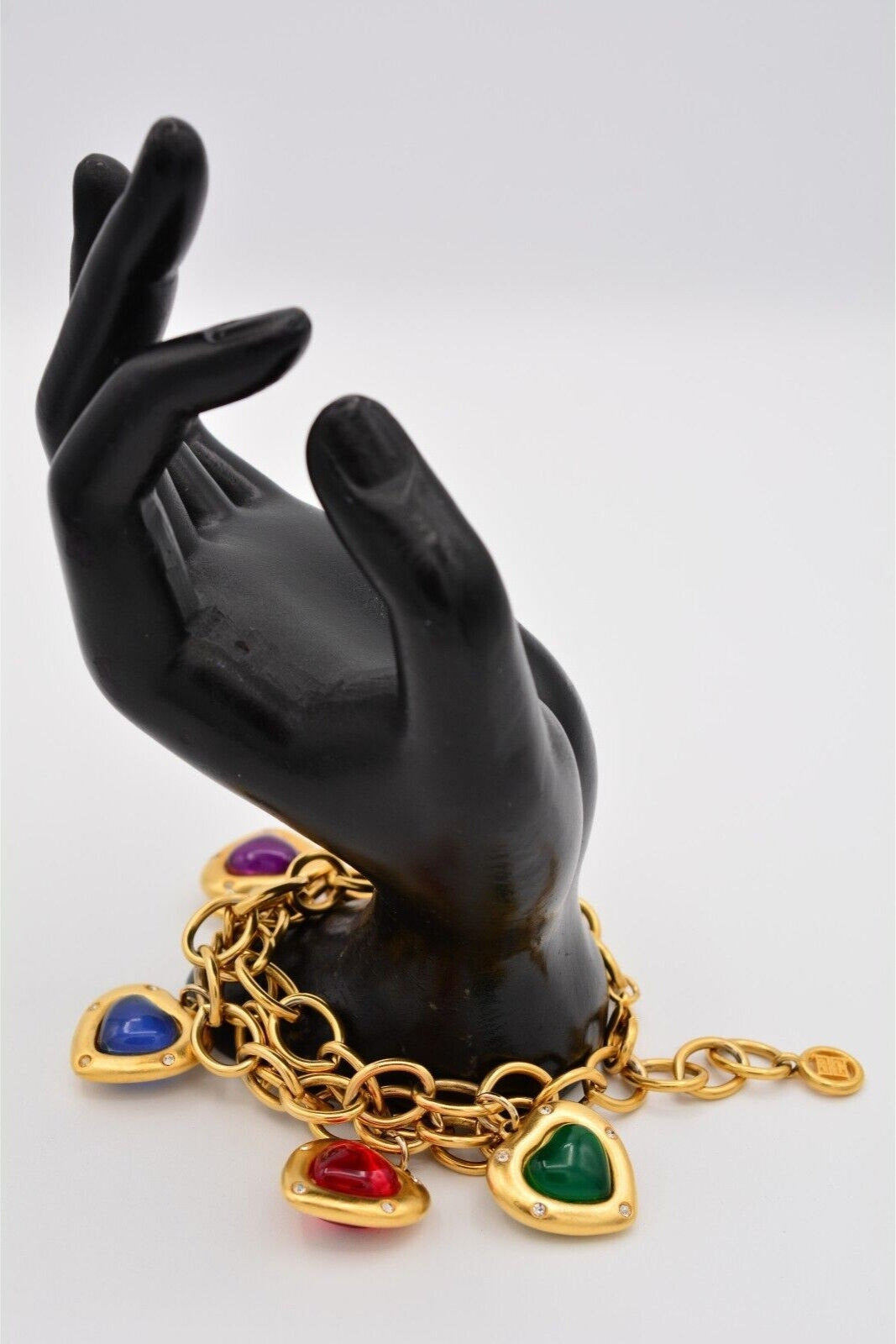 Gold charm bracelet with multiple heart-shaped pendants in red, green, purple, and blue, featuring a glossy, puffy design and small embedded crystals, displayed on a black mannequin hand.