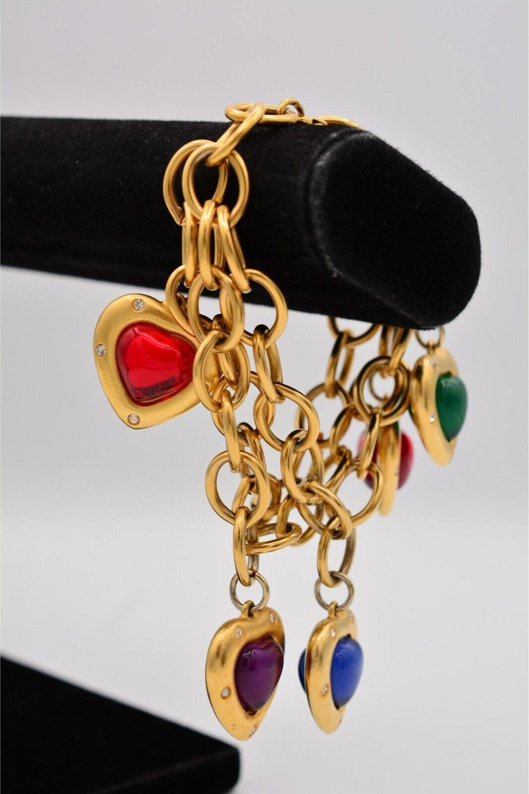 Gold charm bracelet with multiple heart-shaped pendants in red, green, purple, and blue, featuring a glossy, puffy design and small embedded crystals, displayed on a black velvet stand.