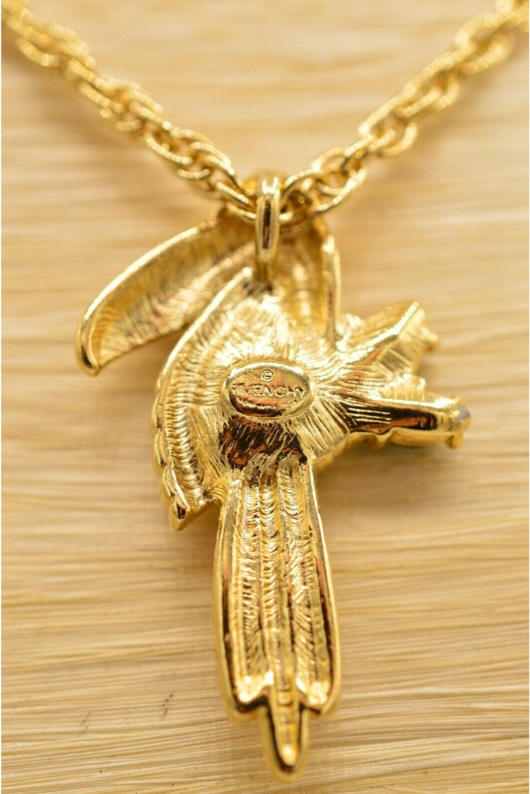 A vintage gold Toucan pendant with colorful red and green crystals, featuring the Givenchy signature on the back, resting on a light wooden neck.
