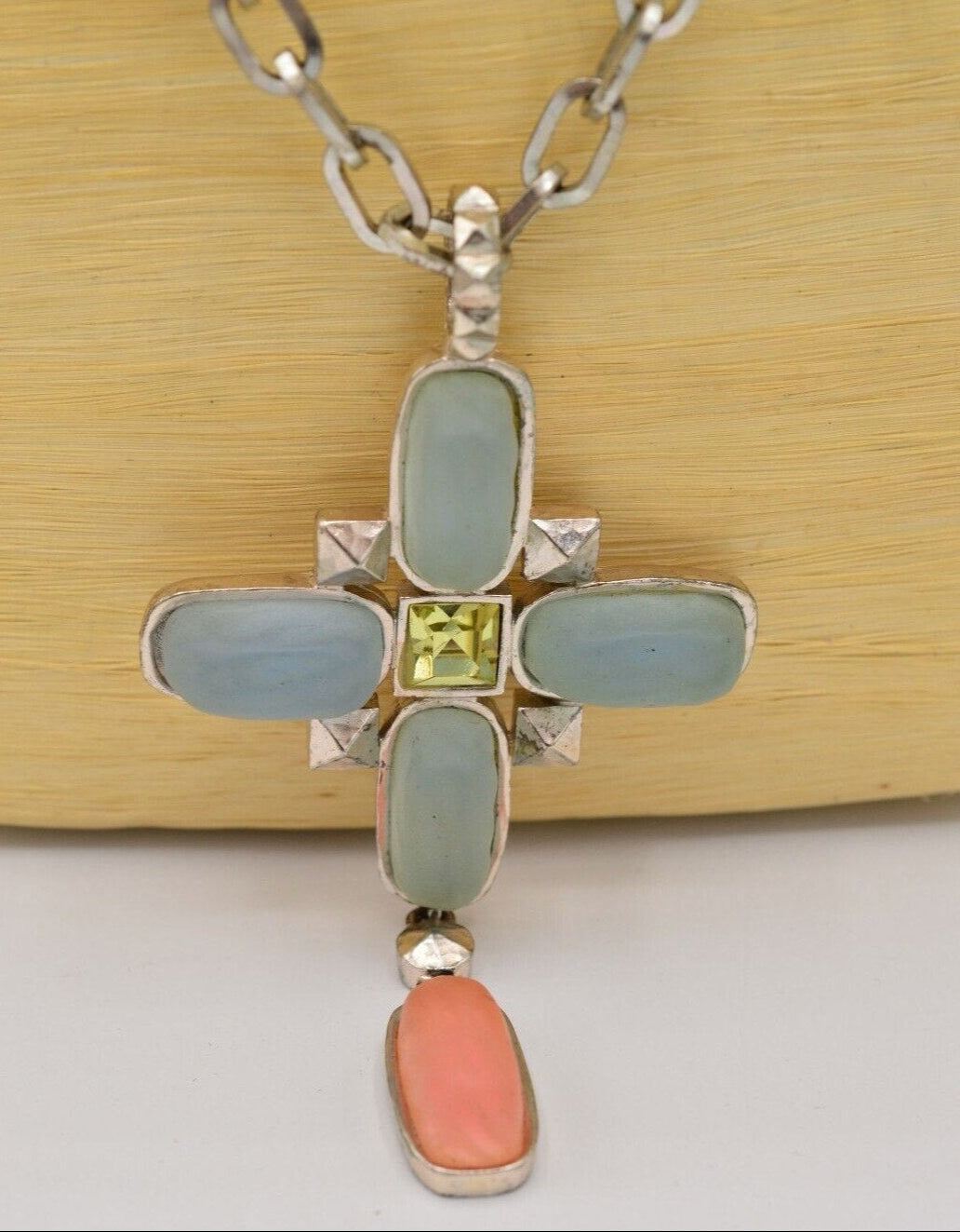 Front view of a vintage Karl Lagerfeld  silver cross pendant with blue and pink frosted glass accents and a vibrant green princess cut rhinestone in the center on a light colored background
