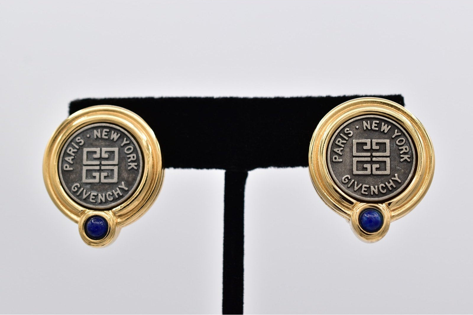 A pair of Givenchy Paris-New York stud earrings in gold-tone metal with a dark gray enamel center, featuring the brand's monogram logo and name, and a small blue gemstone below, displayed on a black T-stand.