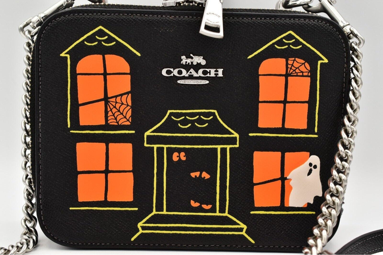A Coach black shoulder bag with a silver chain strap, decorated with an orange and yellow haunted house design and a small white ghost on the front.