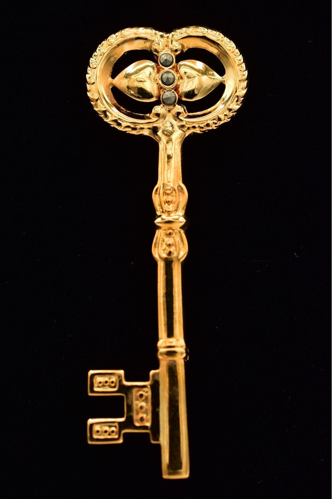 An ornate gold-colored key-shaped brooch or pin with a heart-shaped top and small dark gemstone accents, displayed against a solid black background.