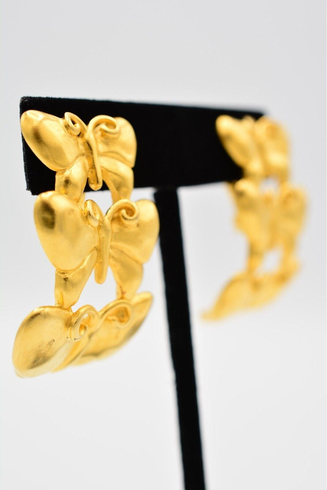 A pair of gold butterfly-shaped earrings displayed on a black jewelry stand against a light gray background.