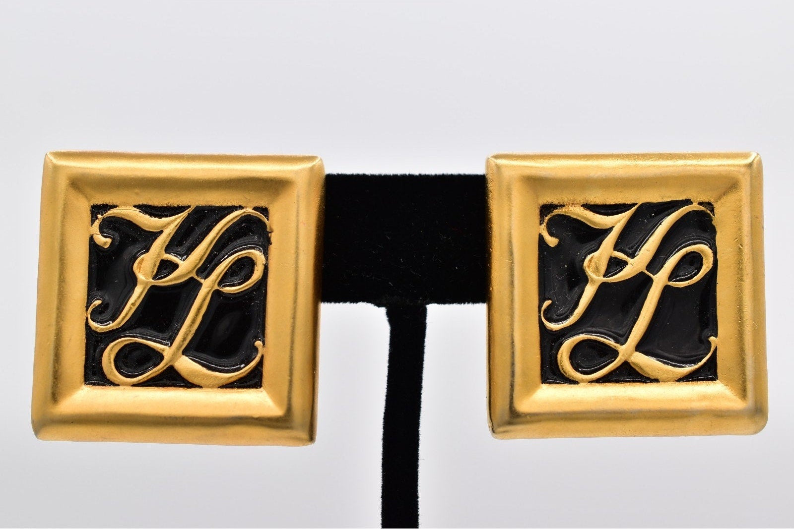 Gold earrings with monogram design on a black band against a light gray background