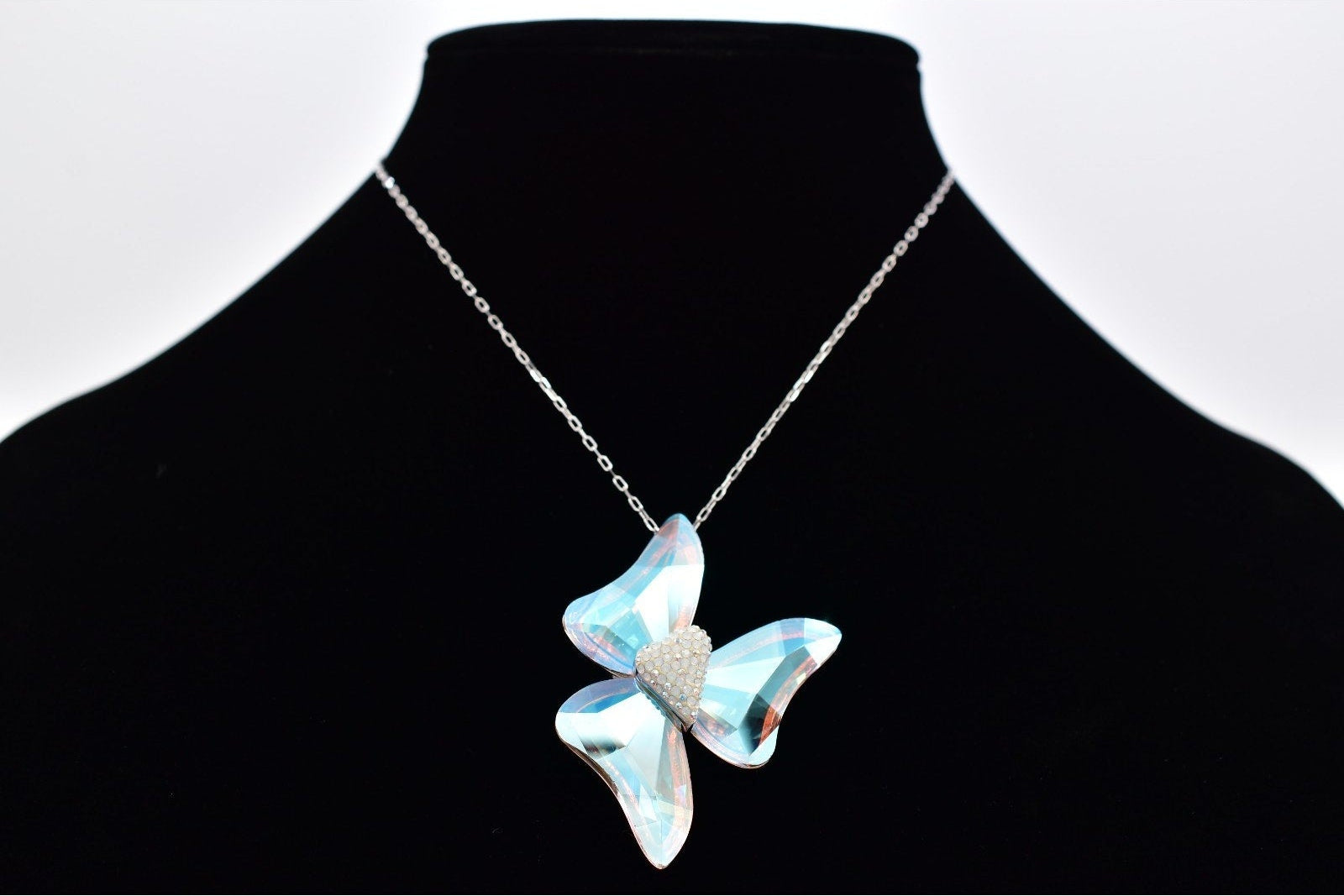 A silver-tone chain necklace with a butterfly-shaped pendant made of iridescent light blue crystals, displayed on a black jewelry bust against a white background.