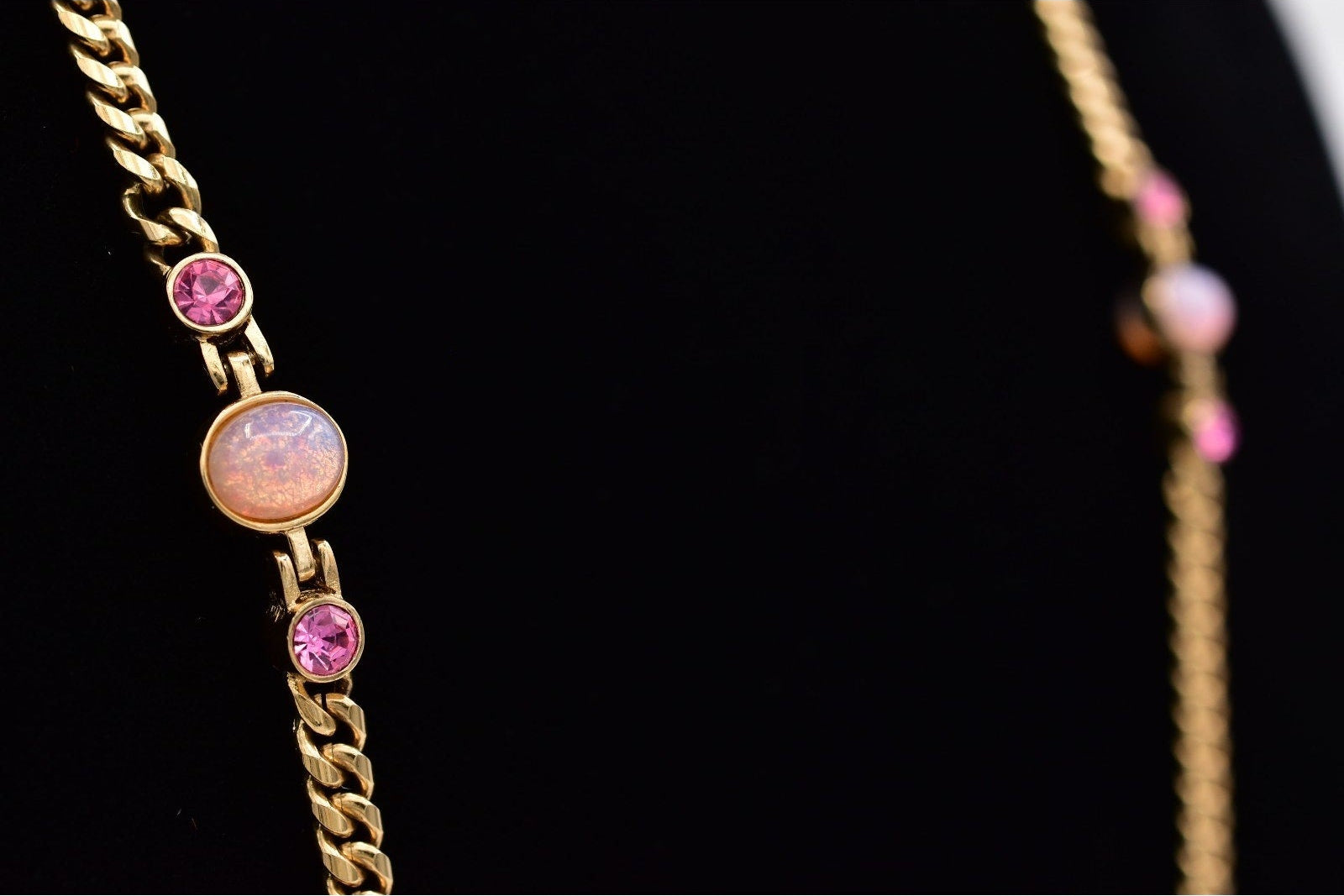 A gold chain necklace featuring a central oval opal-like stone and two round pink gemstones, displayed on a black background.