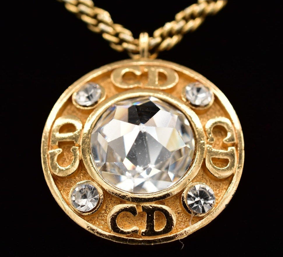 Gold necklace with a circular pendant featuring a large crystal and 'CD' initials on a black background.