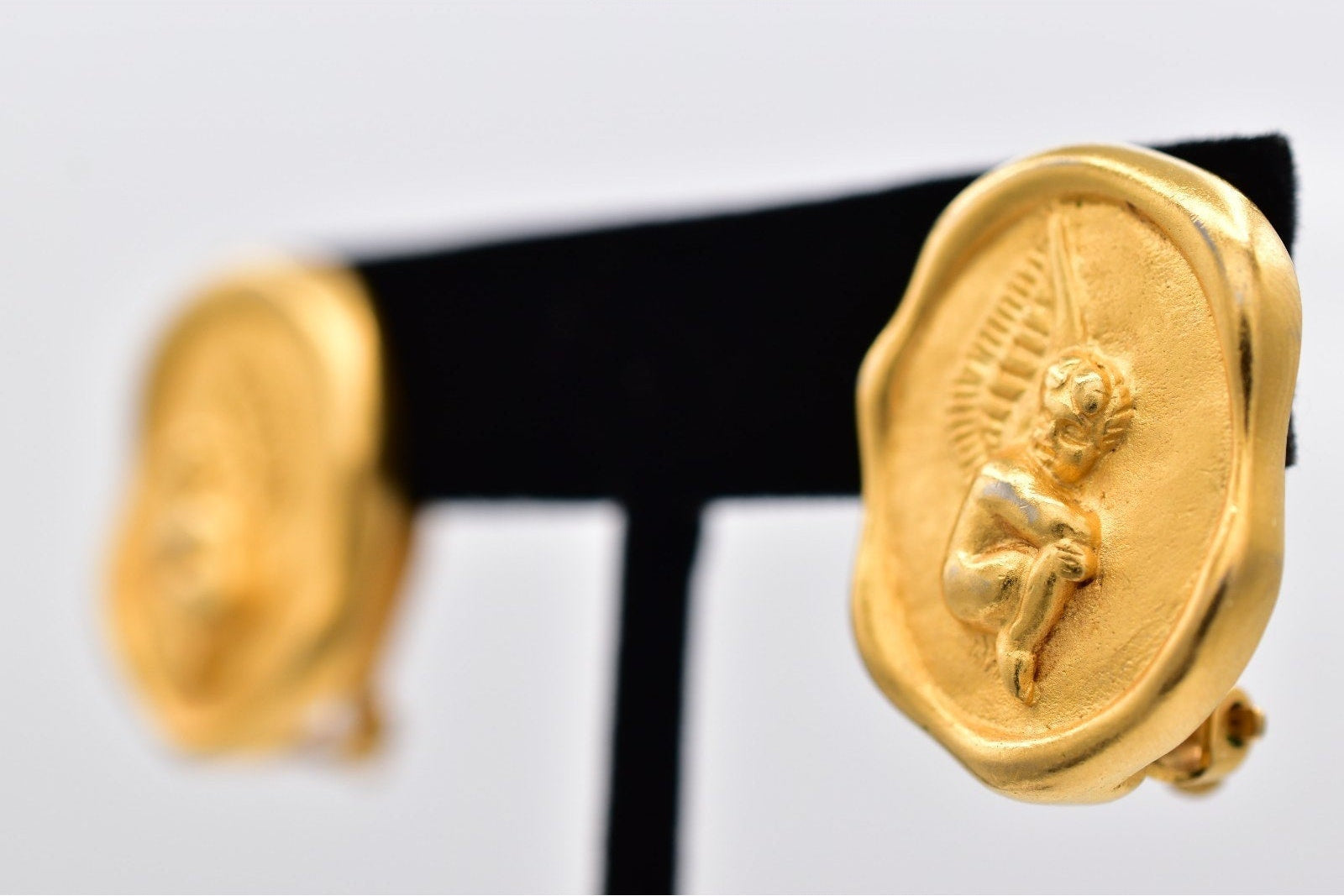 Gold earrings with a cherub design on a black stand against a light gray background