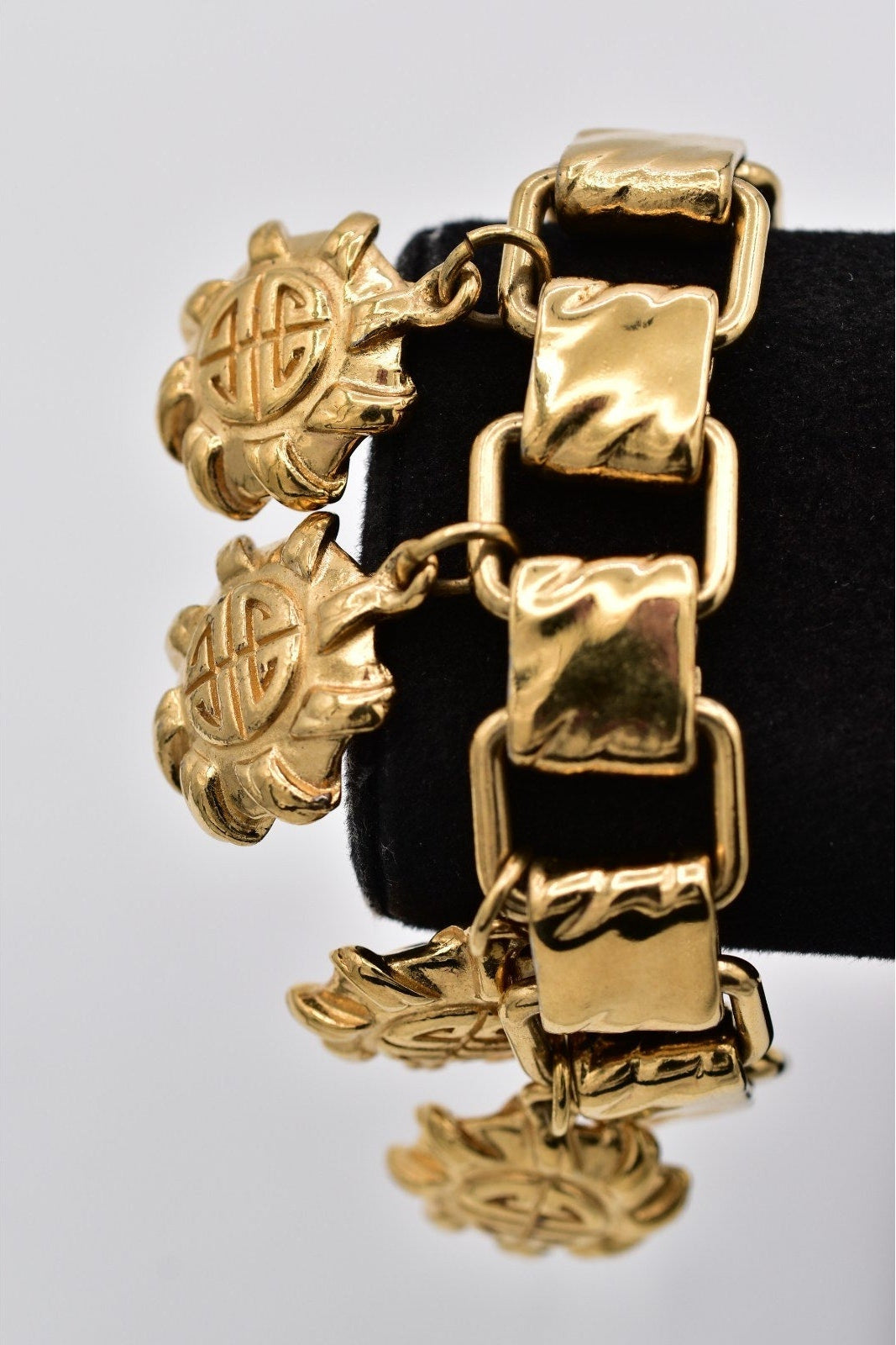 A gold-tone bracelet with textured rectangular links and two dangling sunburst charms featuring a circular emblem, displayed on a black jewelry stand against a light gray background.