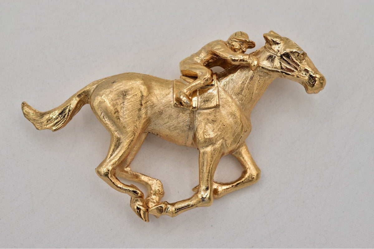 A gold-colored horse and jockey brooch or pin, shown against a neutral light gray background.