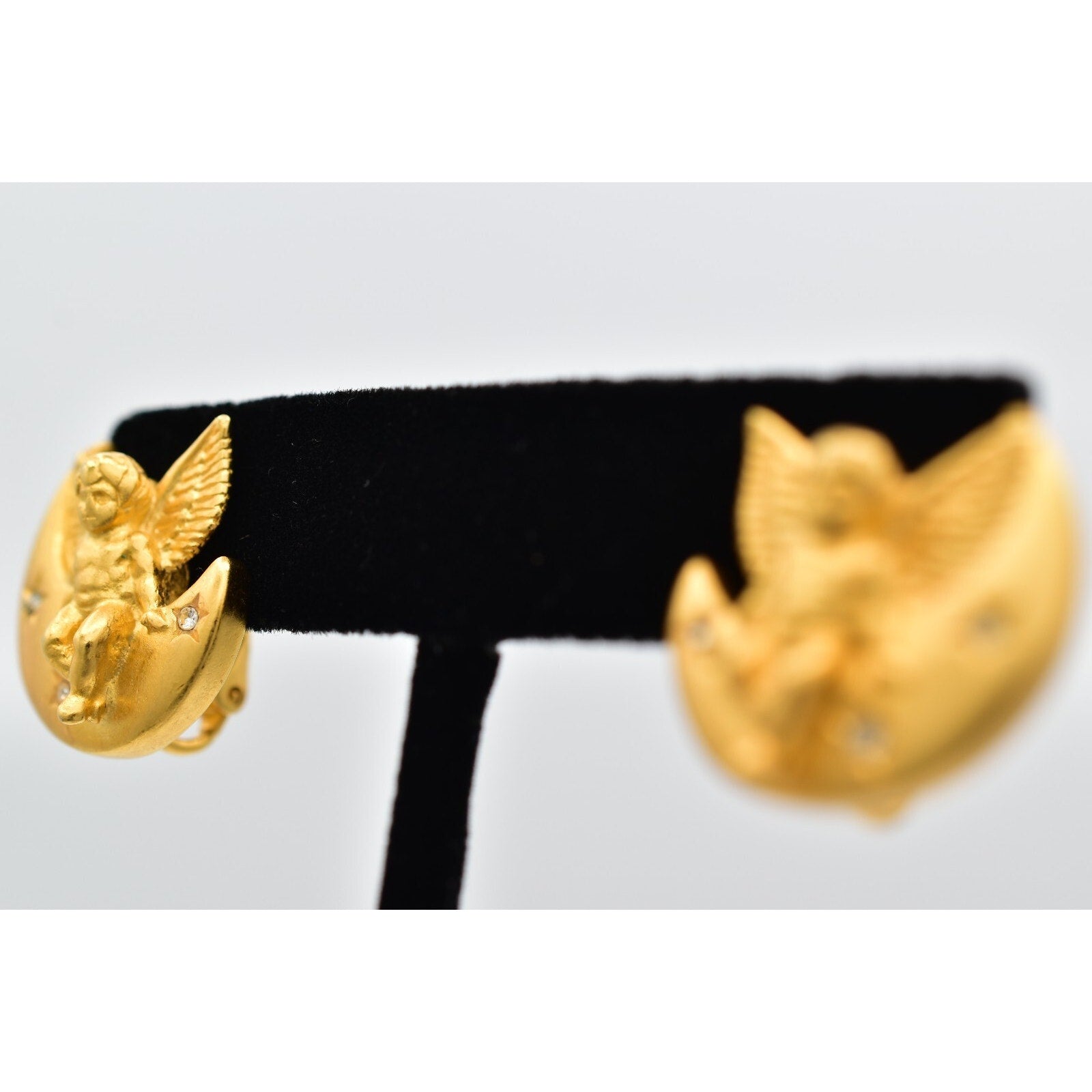 Gold earrings with angel design on a black stand against a light gray background