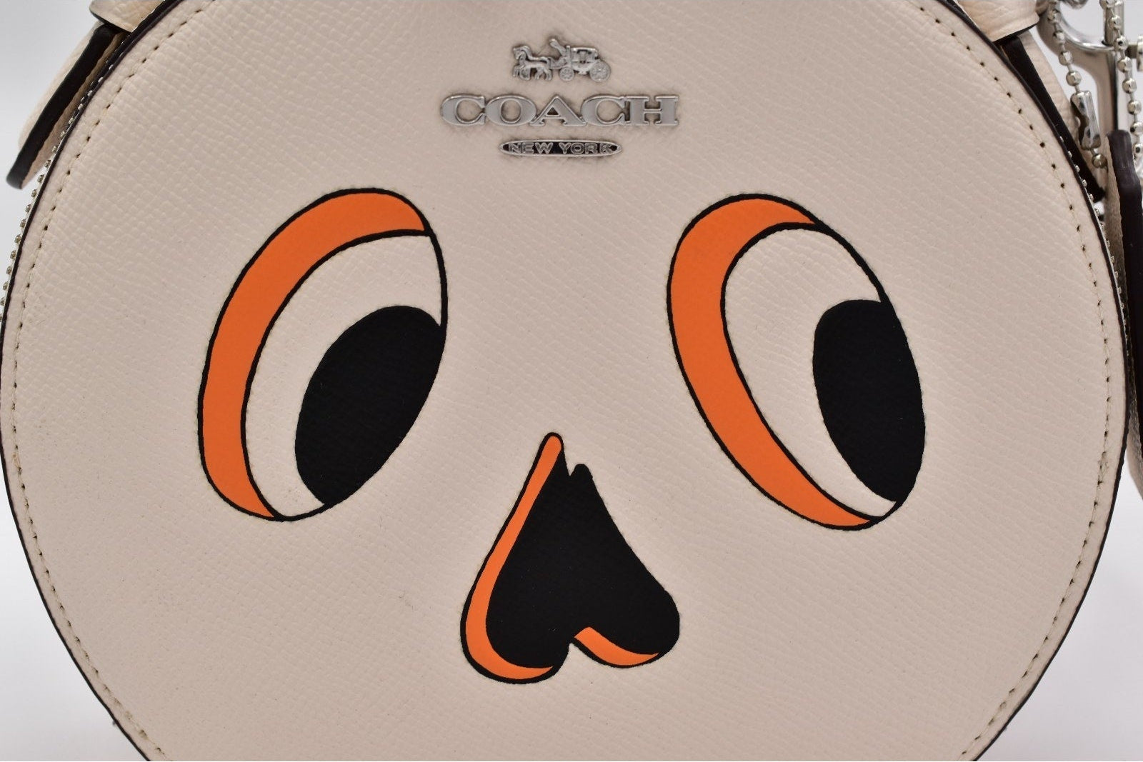 A close-up of a Coach handbag featuring a stylized cartoon face design in orange, black, and white on a light beige textured leather background.