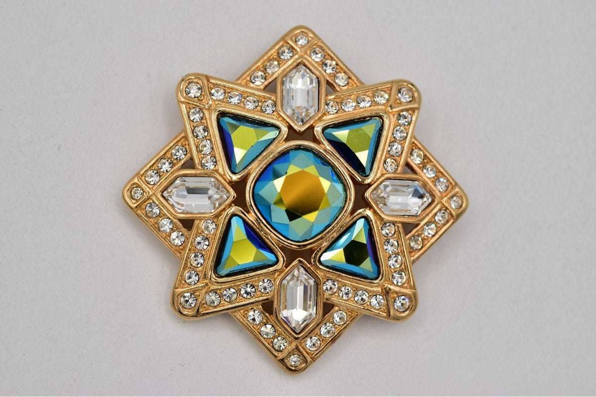 A gold-toned star-shaped brooch featuring iridescent blue and green faceted crystals, clear rhinestones, and a central blue and gold oval stone, set against a plain light gray background.