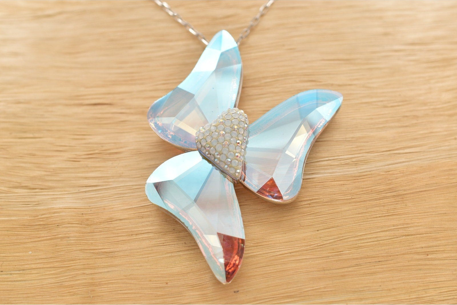 A butterfly-shaped pendant necklace made of iridescent light blue and white rhinestones, with a textured silver heart in the center, resting on a light wood surface.