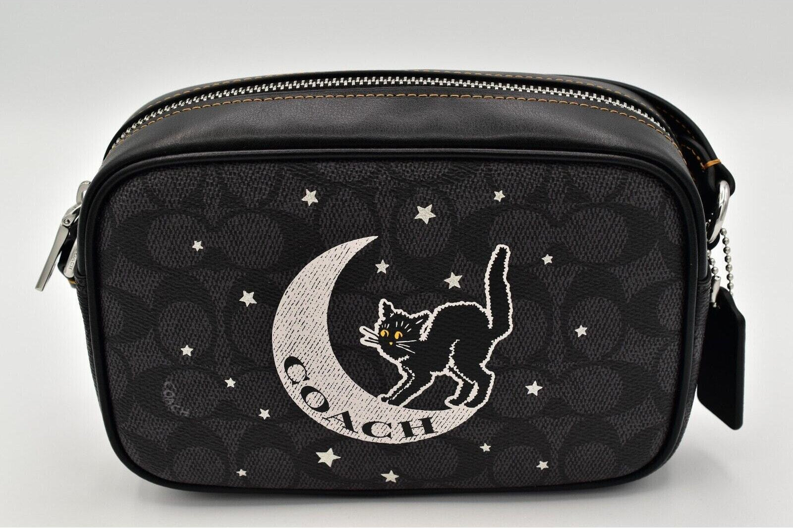 A black Coach crossbody bag featuring the signature monogram pattern, a large embroidered black cat on a silver crescent moon with the Coach logo, and small silver stars.