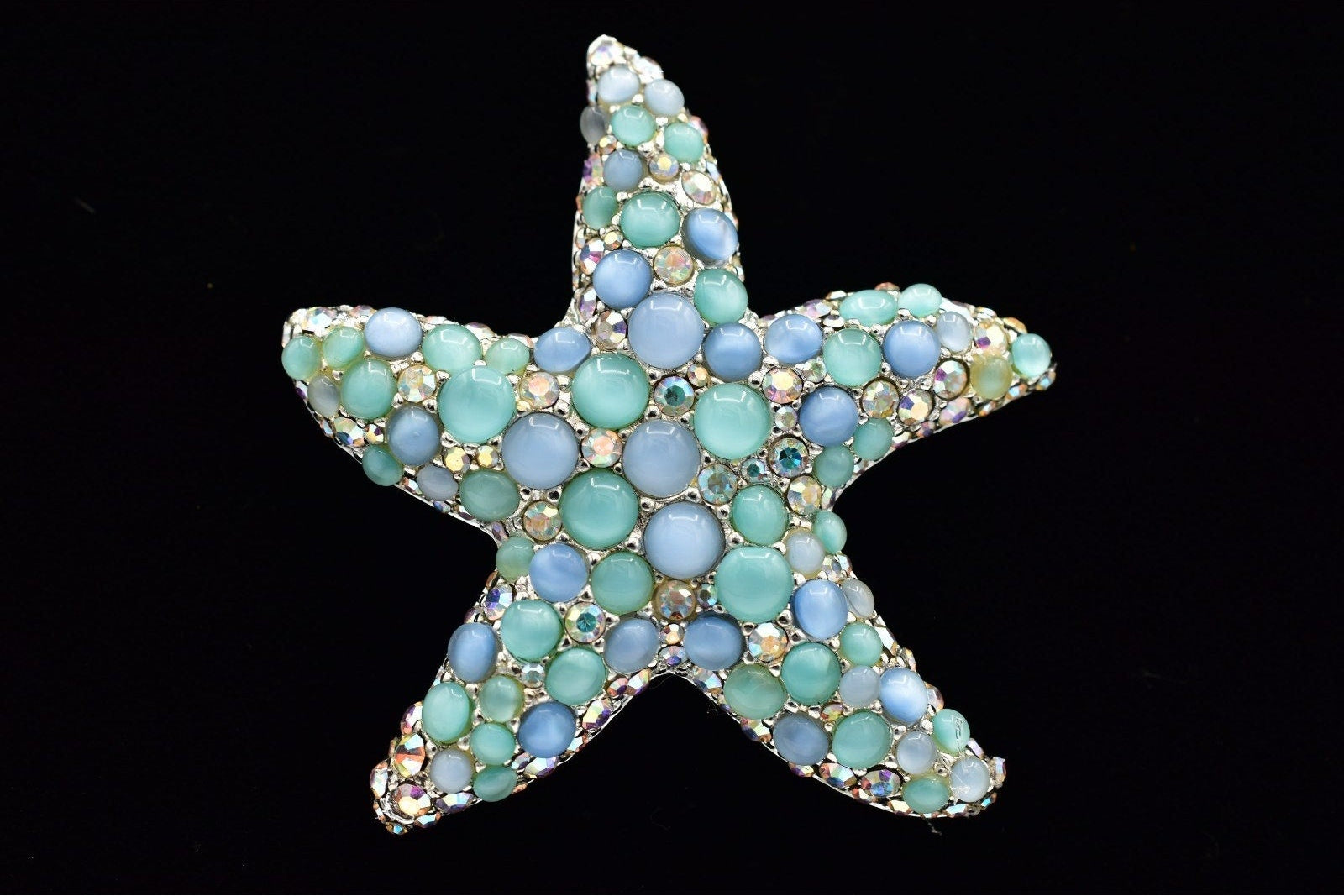 A starfish-shaped brooch embellished with small round turquoise, light blue, and pale aqua stones in a gold-tone setting, presented on a solid black background.