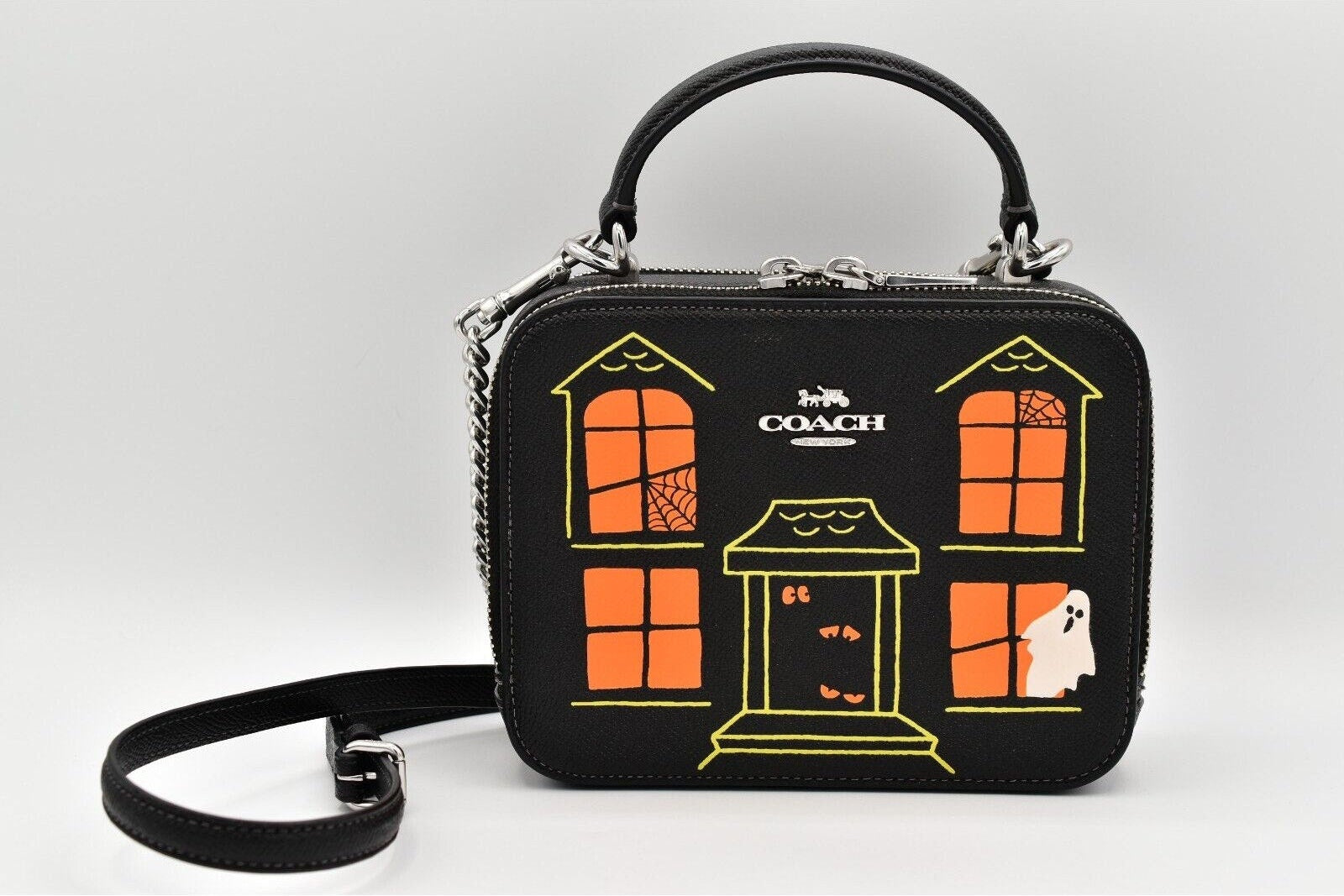 A Coach handbag with a black body and a Halloween-themed house graphic in orange and yellow, featuring a top handle, shoulder strap, and silver hardware.