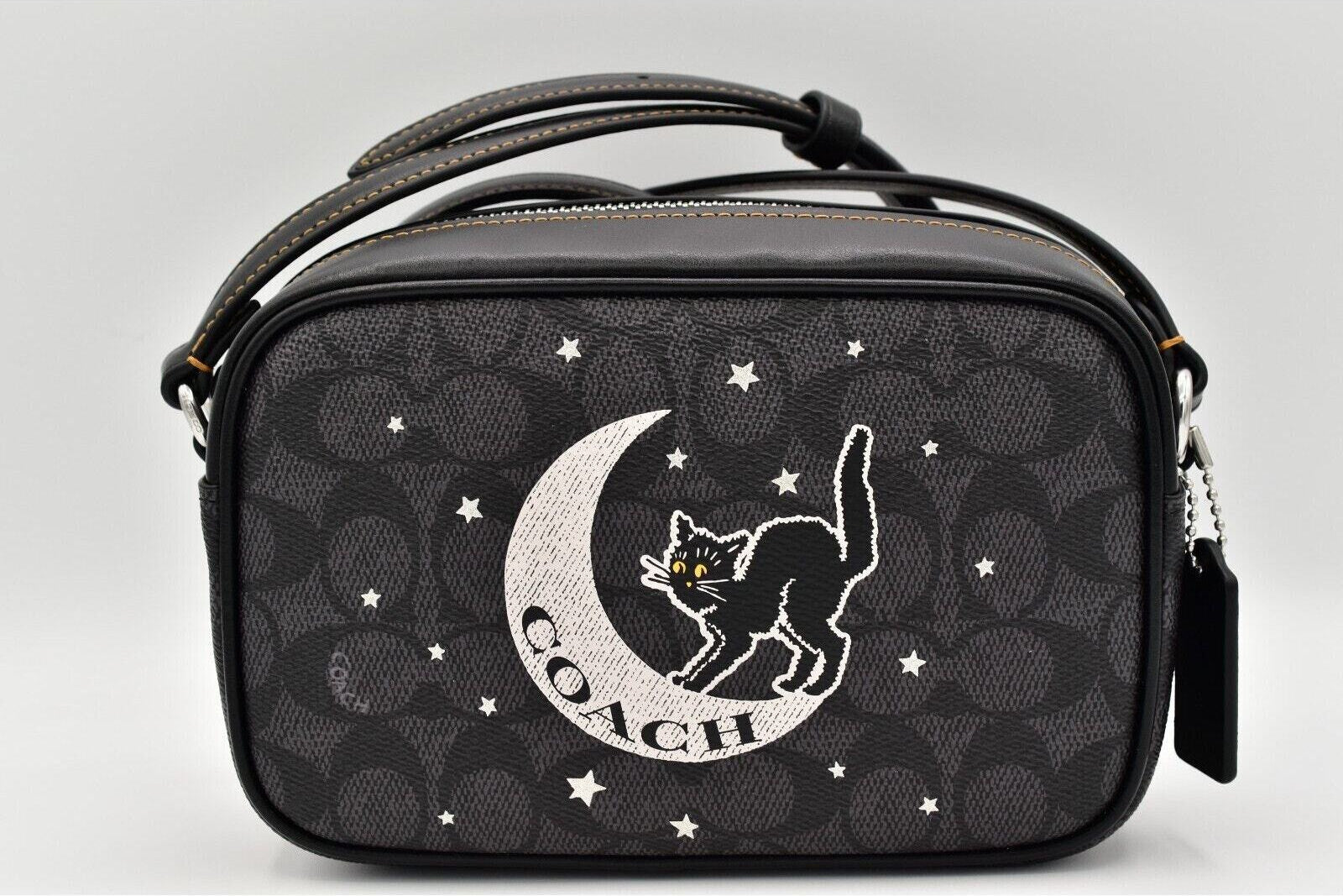 A Coach crossbody bag in black signature monogram canvas with a large white embroidered cat on a crescent moon and the word 'COACH' on the front, shown against a light gray background.