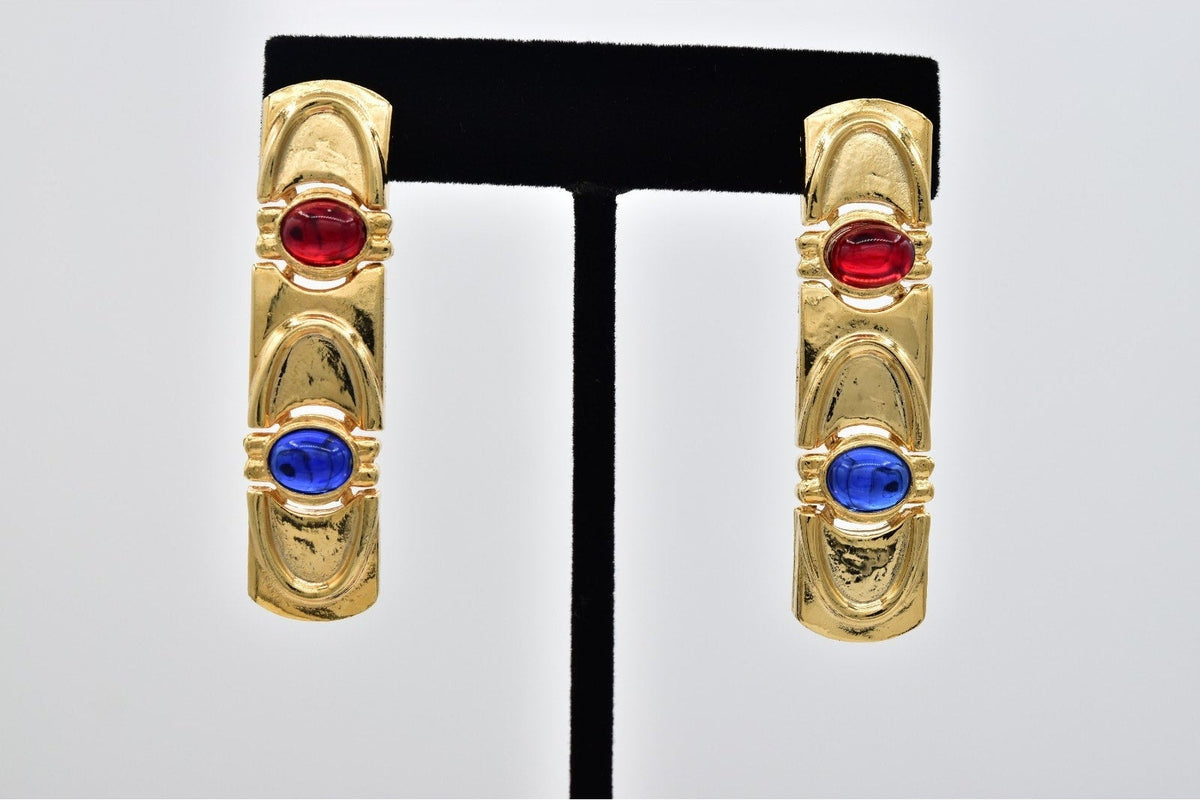 A pair of gold huggie hoop earrings with one red oval gemstone and one blue oval gemstone on each earring, displayed on a black T-stand against a light gray background.