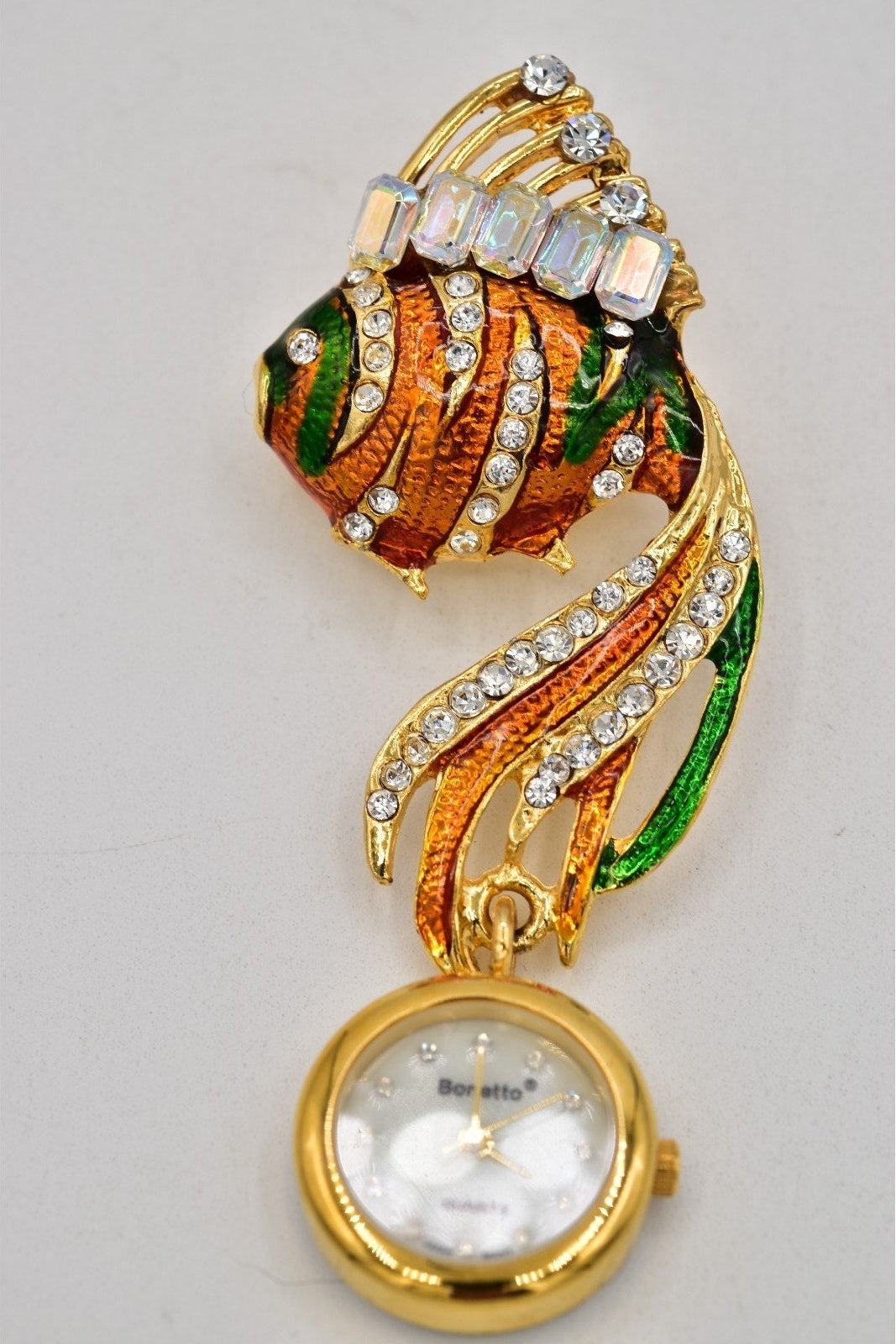 A gold-plated fish-shaped pendant watch with a white mother-of-pearl dial and clear rhinestone accents, shown against a plain light gray background.