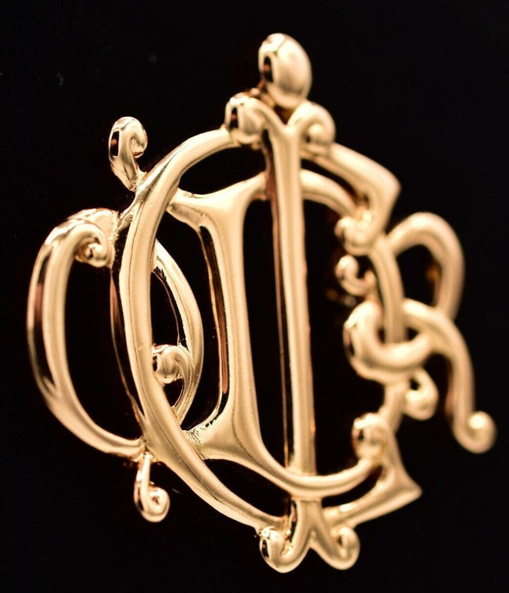 Angled view of a vintage Christian Dior insignia brooch in polished gold tone on a black background. 